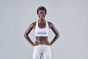 fiducioso giovane africano donna nel gli sport capi di abbigliamento Tenere mani su anca mentre in piedi contro grigio sfondo foto