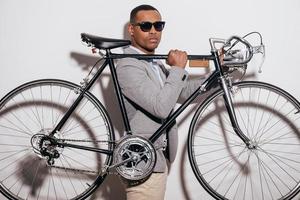 nel il suo proprio stile. fiducioso giovane africano uomo nel occhiali da sole trasporto il suo retrò styled bicicletta su spalla e guardare a telecamera mentre in piedi contro bianca sfondo foto