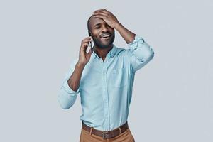 contento giovane africano uomo parlando su il inteligente Telefono e sorridente mentre in piedi contro grigio sfondo foto