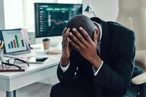 stanco giovane africano uomo nel vestito formale conservazione testa nel mani mentre Lavorando nel il ufficio foto