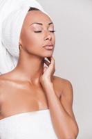 puro bellezza. bellissimo giovane afro americano donna avvolto nel asciugamano conservazione occhi chiuso e toccante sua viso mentre in piedi contro grigio sfondo foto