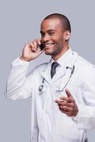 telling bene notizia. contento africano medico parlando su il mobile Telefono e sorridente mentre in piedi contro grigio sfondo foto
