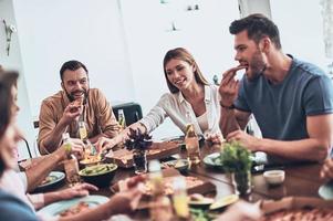 delizioso. gruppo di giovane persone nel casuale capi di abbigliamento mangiare e sorridente mentre avendo un' cena festa in casa foto