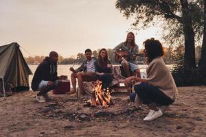 godendo contento momenti. gruppo di giovane persone nel casuale indossare sorridente mentre godendo spiaggia festa vicino il fuoco di bivacco foto