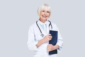 assunzione bene cura di voi. bellissimo anziano donna nel laboratorio cappotto guardare a telecamera e sorridente mentre in piedi contro grigio sfondo foto