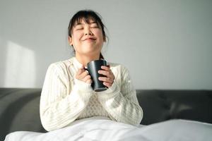 giovane donna potabile caffè nel Camera da letto, Tenere caffè tazza con Due mani foto