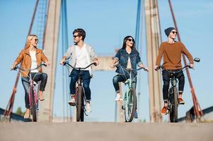 la spesa spensierato tempo insieme. quattro giovane persone equitazione biciclette lungo il ponte e sorridente foto