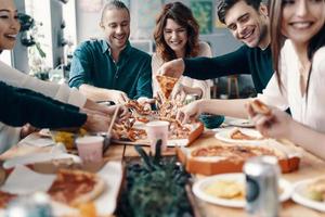 gruppo di giovane persone nel casuale indossare raccolta Pizza e sorridente mentre avendo un' cena festa in casa foto