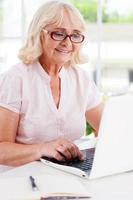 Lavorando a casa. contento anziano donna Lavorando a il il computer portatile e sorridente mentre seduta a il tavolo foto