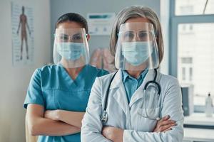 Due donne collaboratori nel medico uniforme e protettivo abbigliamento da lavoro conservazione braccia attraversato e guardare a telecamera mentre Lavorando nel il ospedale foto