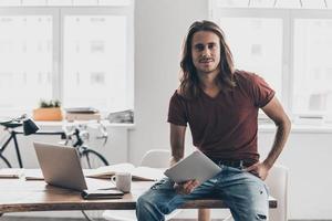 moderno uomo d'affari. bello giovane uomo con lungo capelli Tenere digitale tavoletta e guardare a telecamera con Sorridi mentre pendente a il scrivania nel creativo ufficio foto