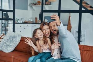 giocoso giovane famiglia sorridente e utilizzando inteligente Telefono per prendere un' autoscatto mentre la spesa tempo a casa foto