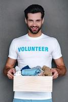 la donazione con piacere. fiducioso giovane uomo nel volontario maglietta Tenere donazione scatola nel il suo mani e guardare a telecamera con Sorridi mentre in piedi contro grigio sfondo foto