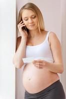 chiamata medico per consultazione. bellissimo incinta donna pendente a il parete e parlando su il mobile Telefono mentre guardare a il carta foto