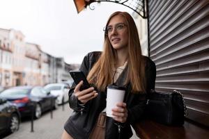 giovane donna con un' tazza di caffè di un' mobile Telefono con un' Sorridi sembra in il distanza contro il fondale di il città foto