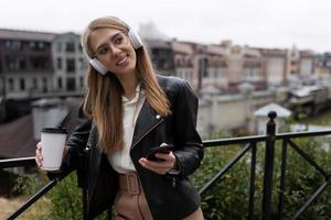 ritratto di un' allegro e contento giovane donna con un' mobile Telefono e un' tazza di caffè nel sua mani ascoltando per musica attraverso sopra l'orecchio cuffie con un' Sorridi su sua viso foto