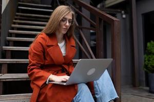 ritratto di un' forte femmina alunno nel un' cappotto seduta su il le scale con un' il computer portatile su sua giro foto