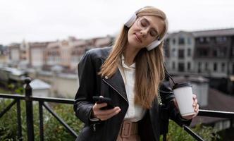 giovane bionda donna per mobile Telefono mani e un' bicchiere di caffè ascolta per musica nel cuffie chiusura sua occhi con piacere su il sfondo di il urbano paesaggio foto