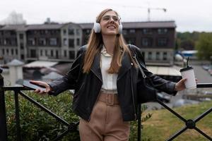 un' giovane donna con un' tazza di caffè nel sua mani gode ascoltando per musica su cuffie contro il fondale di un' città panorama foto