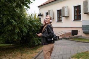 allegro cattivo giovane bionda donna passeggiate in giro il città con un' mobile Telefono e ascolta per musica nel cuffie mentre danza foto