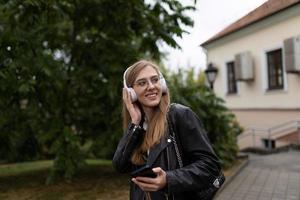 sorridente giovane femmina alunno ascoltando per musica con cuffie su il sfondo di il urbano paesaggio foto