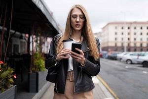 giovane donna digitando un' Messaggio su un' mobile Telefono con un' tazza di caffè nel sua mani contro il fondale di il urbano paesaggio Il prossimo per un' bar foto