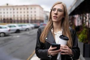 un' giovane ragazza con un' mobile Telefono nel sua mani e un' bicchiere di caffè è guardare per un' modo nel il città foto