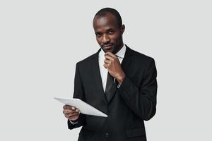 riuscito giovane africano uomo nel vestito formale Lavorando utilizzando digitale tavoletta mentre in piedi contro grigio sfondo foto