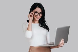 bellissima giovane donna nel inteligente casuale indossare Tenere il computer portatile e regolazione sua occhiali mentre in piedi contro grigio sfondo foto