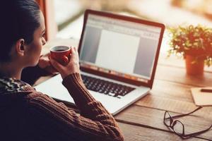 mattina ispirazione. avvicinamento Immagine di pensieroso giovane donna Tenere caffè tazza e guardare lontano mentre seduta a sua Lavorando posto a il ruvido di legno tavolo foto