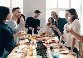 grande festa. gruppo di giovane persone nel casuale indossare mangiare Pizza e sorridente mentre avendo un' cena festa in casa foto