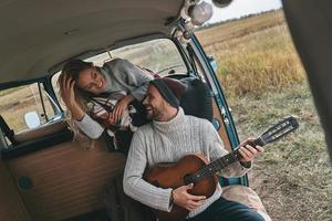 giocando con il suo cuore. bello giovane uomo giocando chitarra per il suo bellissimo fidanzata mentre seduta nel retrò stile mini furgone foto