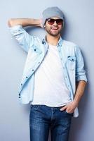 giovane e freddo. bello giovane elegante uomo nel occhiali da sole e cappello guardare a telecamera e sorridente mentre in piedi contro grigio sfondo foto