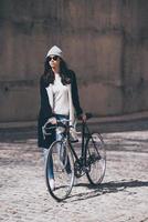 godendo il urbano stile di vita. pieno lunghezza di bellissimo giovane donna nel occhiali da sole rotolamento sua bicicletta e guardare lontano mentre a piedi all'aperto foto