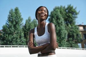 bellissimo giovane africano donna nel gli sport capi di abbigliamento conservazione braccia attraversato e sorridente mentre in piedi all'aperto foto