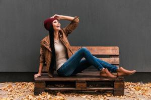 spensierato e così contento. bellissimo giovane donna seduta su il di legno pallet e sorridente con caduto le foglie posa in giro sua foto