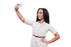 autoscatto volta. bellissimo giovane africano donna fabbricazione autoscatto di sua inteligente Telefono e sorridente mentre in piedi contro bianca sfondo foto