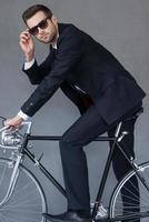 fabbricazione attività commerciale Guarda Buona. bello giovane uomo d'affari regolazione il suo occhiali e guardare a telecamera mentre seduta su bicicletta contro grigio sfondo foto