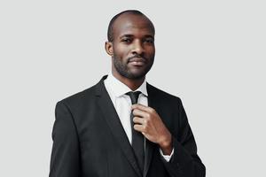 affascinante giovane africano uomo nel vestito formale guardare a telecamera e regolazione cravatta mentre in piedi contro grigio sfondo foto