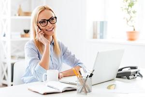 bene attività commerciale parlare. allegro giovane bellissimo donna nel bicchieri parlando su mobile Telefono e sorridente mentre seduta a sua Lavorando posto foto