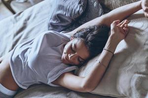 superiore Visualizza di attraente giovane donna conservazione occhi chiuso e sorridente mentre allungamento nel il letto a casa foto