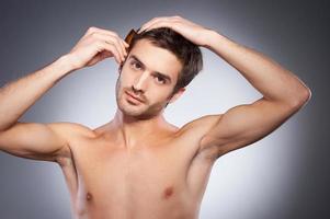 fabbricazione il suo acconciatura. bello giovane barba uomo guardare a telecamera e pettinatura il suo capelli mentre in piedi isolato su grigio sfondo foto