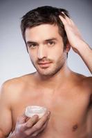 fabbricazione un' affascinante stile. bello giovane A petto nudo uomo toccante il suo capelli con mano mentre Tenere un' contenitore con gel e isolato su grigio sfondo foto