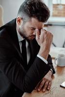 Lavorando tardi. stanco giovane uomo nel vestito formale massaggio naso mentre seduta nel il ufficio foto