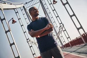 salutare e contento. bello giovane africano uomo nel gli sport capi di abbigliamento sorridente e guardare lontano mentre in piedi su il ponte all'aperto foto