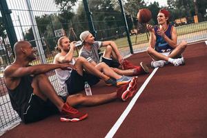 condivisione stesso hobby. gruppo di giovane uomini nel gli sport capi di abbigliamento sorridente mentre seduta su il pallacanestro campo all'aperto foto