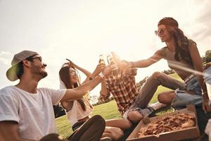 gruppo di giovane sorridente persone nel casuale indossare tostatura con birra bottiglie mentre godendo picnic all'aperto foto