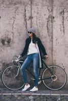 amorevole sua nuovo stile. bellissimo giovane donna nel occhiali da sole Tenere mani su sua bicicletta e guardare lontano mentre in piedi contro calcestruzzo parete all'aperto foto