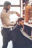 barbiere a opera. lato Visualizza di giovane barbuto uomo ottenere taglio di capelli di parrucchiere mentre seduta nel sedia a barbiere foto