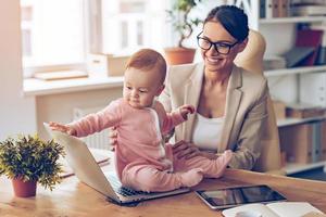 mammina esso è tempo per un' rompere allegro giovane bellissimo donna d'affari guardare a sua bambino ragazza con Sorridi mentre seduta a sua Lavorando posto foto